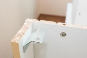 Chipped MDF furniture corner exposing compressed wood fibers beside a metal bracket.